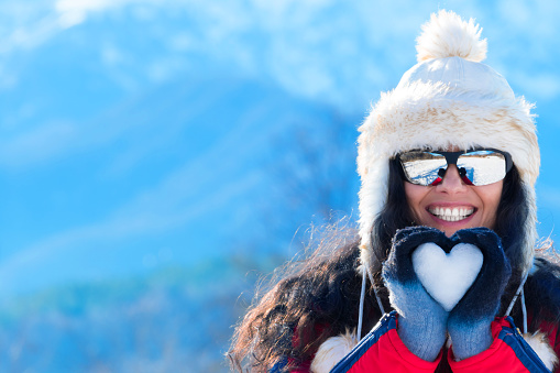 Smiling young woman with long brown hair and sunglasess making snow heart in winter mountain. Wear white fluffy hat, blue-black gloves and red jacket. As background is the snow mountain.