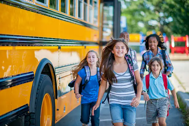 A multi-ethnic group of elementary age children are running from the bus to class. They are smiling and looking at the camera.