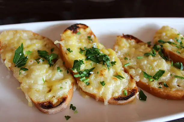Photo showing slices of grilled garlic bread, topped with melted cheese and fresh herbs, including parsley.  The garlic bread was served on a long white plate in a Spanish restaurant, as part of a 'tapas' meal.