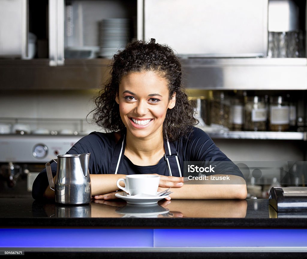 Coffee shop employee Friendly afro american girl standing behind the bar counter and serving coffee in coffee shop, smiling at camera. African Ethnicity Stock Photo