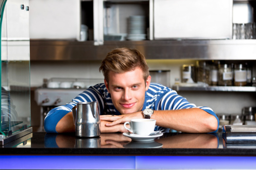 Friendly young man standing behind the bar counter and serving coffee in coffee shop, smiling at camera.