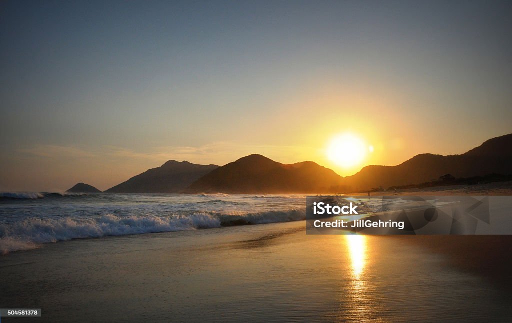 Sunset on  travel destination beach in exotic Brazil Beautiful Brazilian beach in Rio de Janeiro. Sun sets behind a mountain ridge on a October evening in this tropical paradise in South America. Way in the distance a couple stands close together. The sun reflects on the wet sand in the foreground. Waves crashing on the left side of the frame. Beach Stock Photo