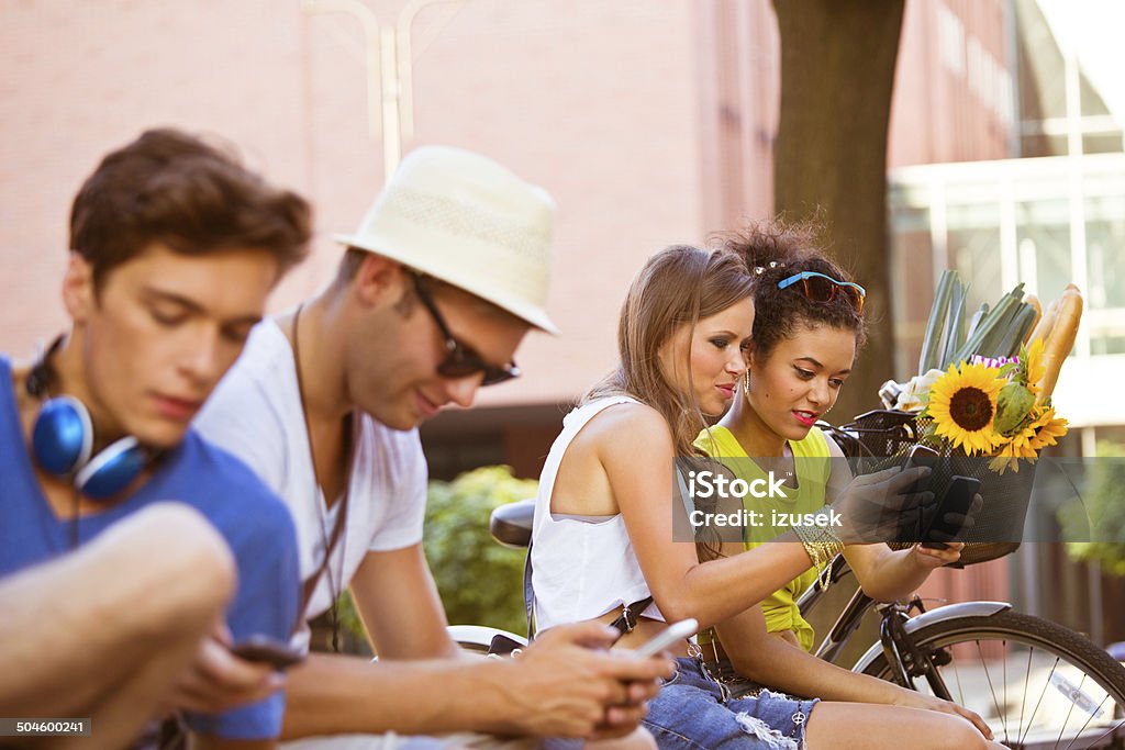 Urban young people Four young urban people sitting outdoors and using their smart phones. Focus on two girls sitting in the background. 20-24 Years Stock Photo