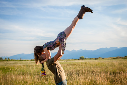 A handsome young man lifting up his girlfriend. She is holding flowers and wearing cowboy boots. A grassy field, mountains in the distance, and blue sky make a pleasant countryside backdrop.