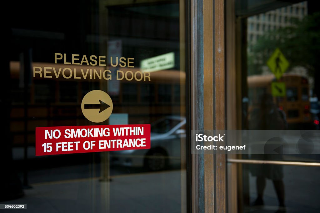 No Smoking Zone A caution notice prohibiting smoking inside 15 feet area of the building entrance. Request to use the revolving door is also seen on the black glass door of the building. Alertness Stock Photo