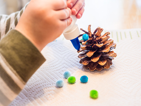 closeup of child's hands as he glues pom poms on a pinecone