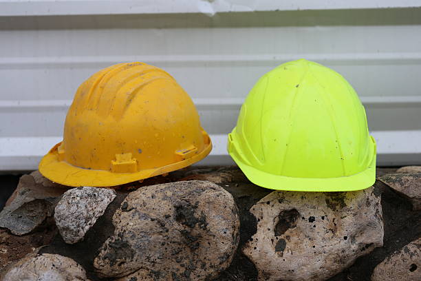 Two Work Helmets Work helmets of a construction worker on a rock fence in the construction site. Yellow and orange wet safety helmets of a construction worker in front of a corrugated iron fence in a rainy day. corrugated metal fence ideas stock pictures, royalty-free photos & images