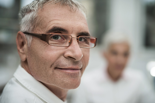 Close up of a smiling mature doctor looking at the camera. There are people in the background.