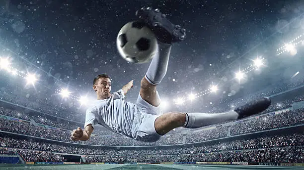 A male soccer player makes a dramatic play by jumping horizontally. He attempts to kick the ball with his feet. The stadium is dark behind him. Only the lights of the stadium shine brightly, creating a halo effect around the bulbs.