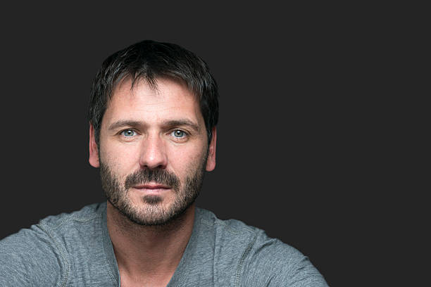 A attractive serious man with black hair and beard looking directly at the camera. His head and shoulders are on the left side and there's some copy space on the right side.