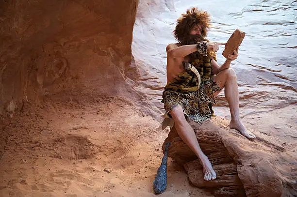 Caveman sitting using touchscreen stone tablet outdoors in a weathered rock cave