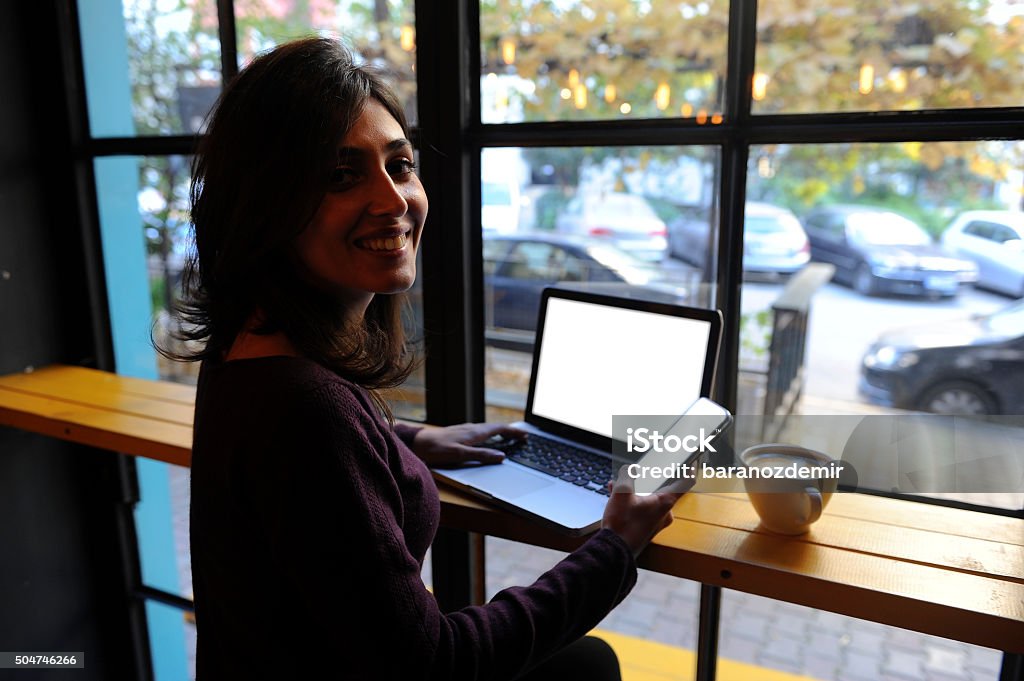 Young woman working in a coffee shop Smiling young woman sitting in modern coffee shop, working with her phone and laptop computer. 20-29 Years Stock Photo