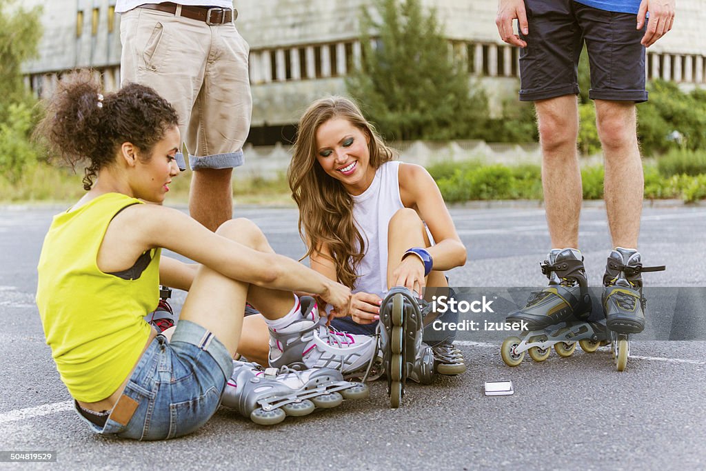 Young people on rollerblades Two young women sitting on tarmac and tying rollerblades with guys standing behind them. 20-24 Years Stock Photo