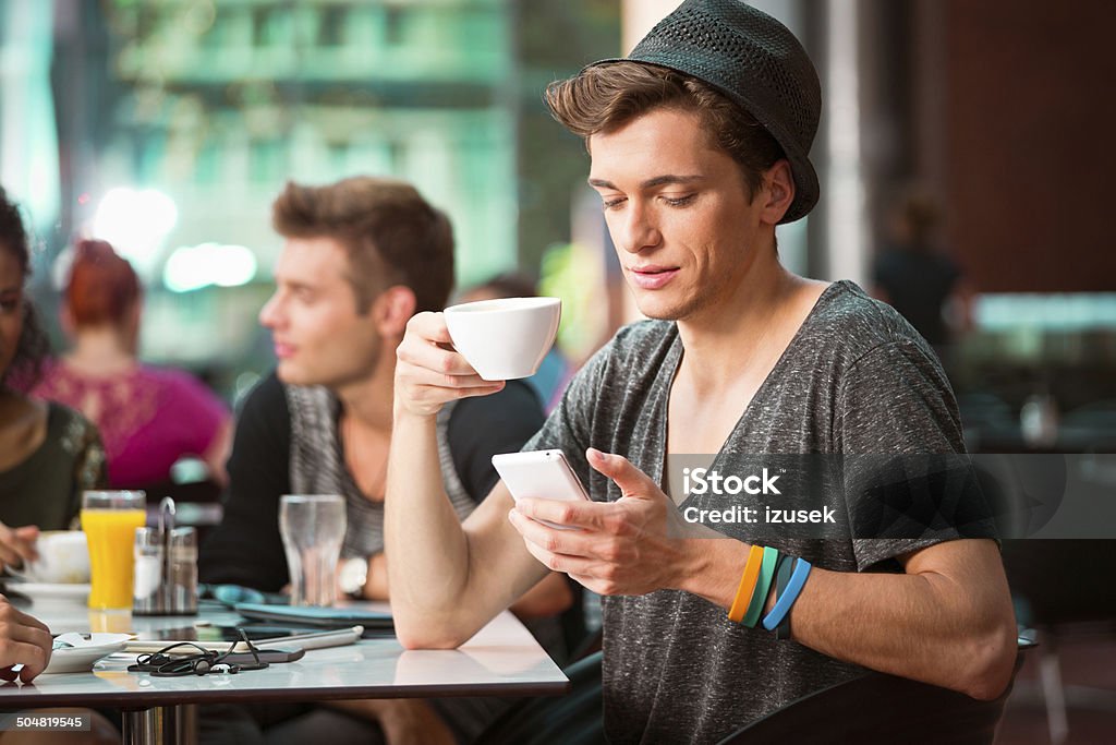 Young man on phone Cheerful young man sitting in coffee shop, holding cup of coffee and texting on phone. People in the background. 20-24 Years Stock Photo