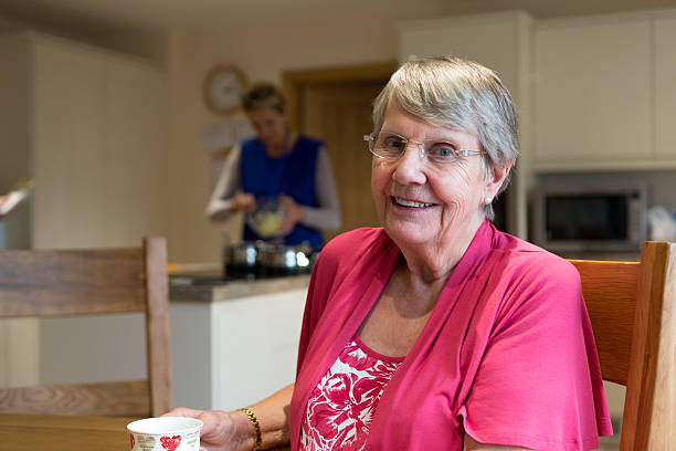 Dinner won't be long now! Elderly woman sitting at her kitchen table waiting for her lunch. She is looking at the camera smiling. Home help is preparing her dinner in the back ground. meals on wheels photos stock pictures, royalty-free photos & images