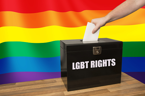 Hand placing a vote in a ballot box on LGBT Rights with a rainbow flag in the background.