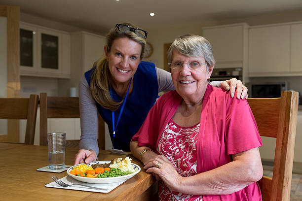 Lunch is served! Community care worker serves up lunch to an elderly woman sitting at her kitchen table. meals on wheels photos stock pictures, royalty-free photos & images