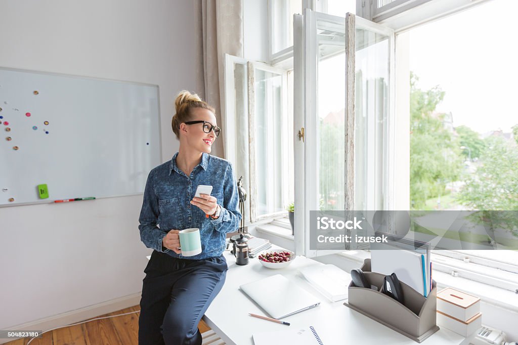 Coffee break, woman sitting on the desk, using smart phone Coffee break in an office. Woman wearing jeans shirt and nerd glasses holding a cup of coffee and using a smart phone, looking through the window. Freelance Work Stock Photo