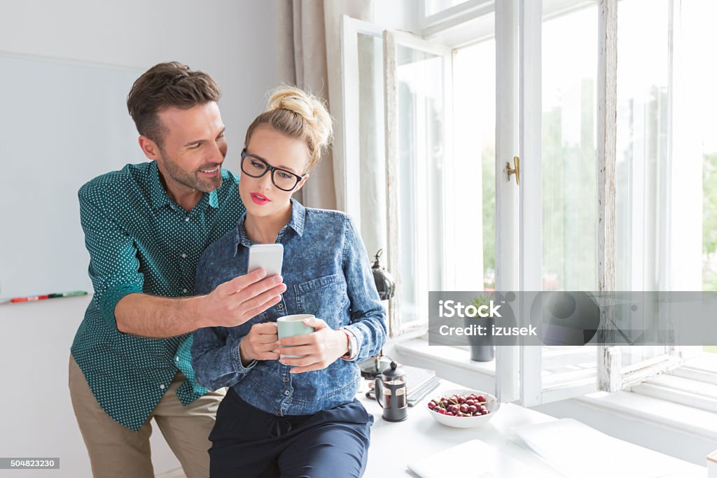Friends using a smart phone together in an office Friends using a smart phone together in a home office, man holding phone and woman holding cup of coffee.  Coffee - Drink Stock Photo