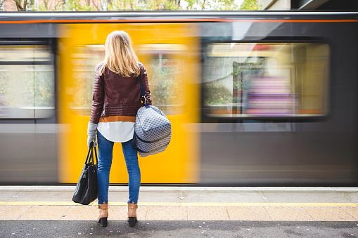 Rear view of a young hipster style woman holding a couple of bags while waiting for the train. A yellow and grey train can be seen moving in front of her in a blur.