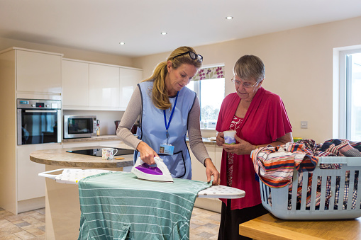 Care worker making a home visit. Female carer is ironing in the kitchen to help an elderly woman. The elderly woman is standing behind her chatting.