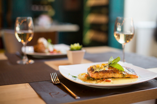 Close-up shot of unrecognizable woman eating salmon steak with vegetables and potato croquette. Her hands cutting the salmon with table knife.