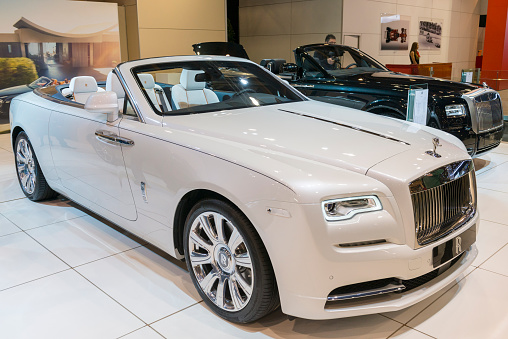 Brussels, Belgium - Januari 12, 2016: White Rolls-Royce Dawn luxury convertible automobile. The car is fitted with a twin-turbo 6.6-litre V12 and luxurious leather and wood interior. The car is on display during the 2016 Brussels Motor Show. The car is displayed on a motor show stand, with lights reflecting off of the body. There are people looking around and other cars on display in the background.