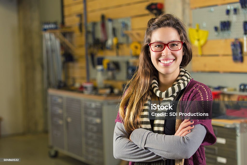 Hipster college student or entrepreneur in makerspace workshop Young adult Caucasian hipster woman is smiling and looking at the camera. College student or artist entrepreneur is standing confidently in makerspace or workshop studio. Variety of woodworking tools and supplies are organized in background. Music Store Stock Photo