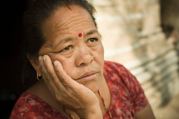 Outdoor image in day time of a sad Asian mature woman holding head and looking up with blank expression. She is a poor peasant and living in the village of India and Nepal. One person, head and shoulders, horizontal composition with selective focus and copy space.