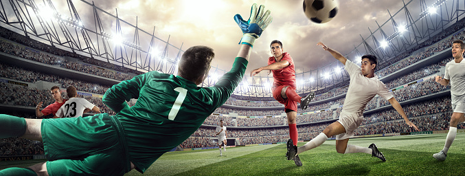A male soccer player kicks a ball and soccer goalie is ready to block it with his hands in a jump on wide angle panoramic image of a outdoor soccer stadium or arena full of spectators under a sunny sky. The image has depth of field with the focus on the foreground part of the pitch. With intentional lensflares. Players are wearing unbranded soccer uniform.