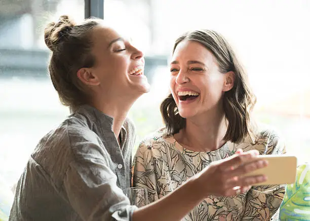 Two female friends with smartphone, smiling. Young woman holding phone, mid adult woman looking at her and laughing