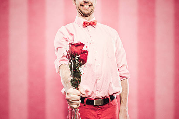 A man in pink and red clothes and a bow tie has a valentine bouquet of roses to give to his love.  Bright pink striped background.  Image for Valentines holiday.  Horizontal image with copy space.