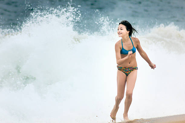 A DSLR photo of a cute 9-year-old caucasian girl enjoying her vacations at the beach. She is playing near the water running awat from a splashing wave. She is wearing a colorful bikini. It is past sunset and the light is very soft.