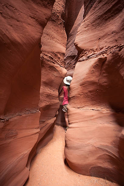 Exploring woman hiking slot canyon Escalante National Monument Utah As the knobby and striated cliff walls close in, a woman with long hair, shorts, t-shirt and hat hikes along the sandy bottom of the wind and water eroded slot canyon and cliffs of Spooky Gulch in the Escalante National Monument, Utah. grand staircase escalante national monument stock pictures, royalty-free photos & images