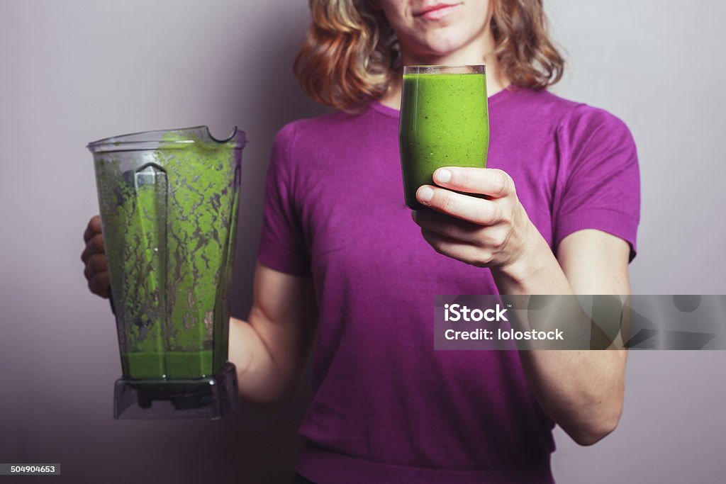 Young woman with green smoothie A young woman in a purple top is holding a blender and a glass with green fruit smoothie Adult Stock Photo