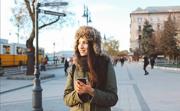 Winter portrait of a young brunette in Budapest. She is wearing a jacket and a Russian style fake fur hat, walking by the river Danube, lit by the evening sun, looking at her smartphone, texting, browsing the web. Smartphone social networking on the street concept.