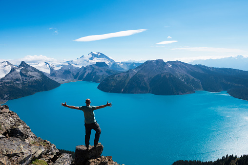 Man celebrating a personal victory by raising his arms at a pristine glacial alpine lake flanked by mountains in the distance.  This photo represents spirituality, religion, victory, triumph, connection, teamwork, inspiration, success, aspirations, conquering adversity, authenticity, winning and several other concepts.