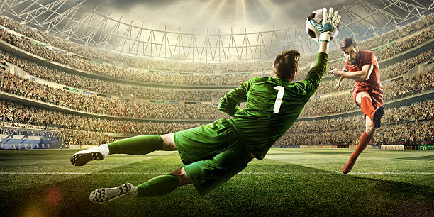 Soccer game moment with goalkeeper A male soccer player kicks a ball and soccer goalie jumping in motion for a ball while defending his gates on wide angle panoramic image of a outdoor soccer stadium or arena full of spectators under a sunny sky. The image has depth of field with the focus on the foreground part of the pitch. With intentional lensflares. Players are wearing unbranded soccer uniform. taking a shot sport stock pictures, royalty-free photos & images