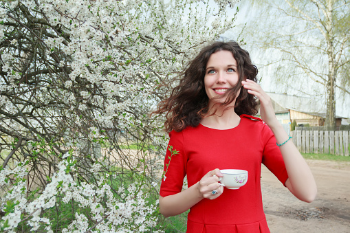 Cheerful young lady in red dress and waving hair in windy spring blooming white fruit garden