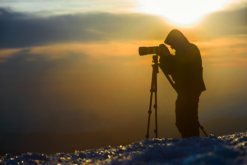 Photographer are making landscape shot in mountains.  Shine snow is in foreground. Silhouette of man is situated against beautiful multicolored sky background.