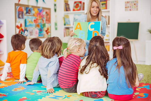 Playful learning Teacher with a group of preschool children in a nursery. The children are sitting on the floor and listening  teacher. Learning letters. In the background we can see a shelf with some, toys, black board and books. View from behind. child care photos stock pictures, royalty-free photos & images