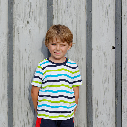 Portrait of happy little cute kid boy with blond hairs and blue eyes against gray wooden background, outdoors. Funny child.
