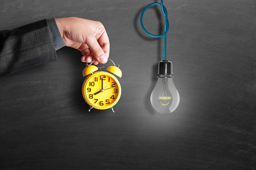 Close-up of a businessman hand holding yellow alarm clock with hanging light bulb on blackboard. Concept of quality work with time management.