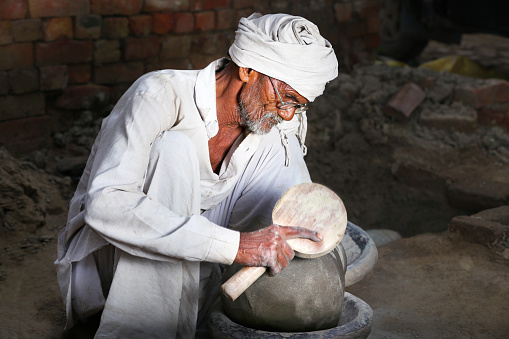Senior Potter of Indian Ethnicity Making Sitting at his home and Making ceramic pots. He is wearing white dhoti-kurta & turban which is traditional clothing for men in north India Portrait using studio lights.