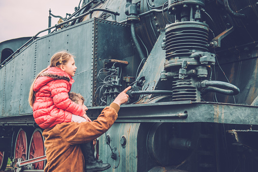 Girl with long brown hair in pink winter jacket riding on her grandfather's shoulders and admiring an old steam locomotive on the Nova Gorica train station, Slovenia, Europe. Side view, close-up. The station  was opened in 1906 in Austrian Empire. Nikon D800, full frame, XXXL.