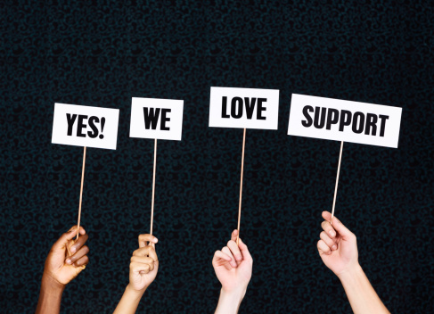 Four hands hold up words saying 'Yes! We love support'  against a darkly patterned background with some copy space. As long as you have support, you can do anything!