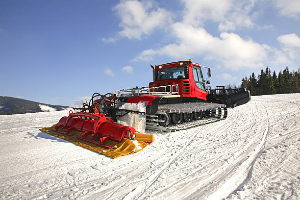 A snow groomer in vibrant red color is actively shaping the ski slope on a mountain, ensuring a smooth surface for skiers. The clear blue sky and rugged terrain add to the picturesque alpine setting.