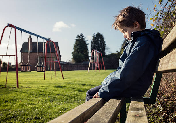 Lonely child sitting on play park playground bench Upset problem child sitting on play park playground bench concept for bullying, depression, child protection or loneliness sulking stock pictures, royalty-free photos & images