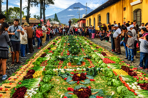 Antigua, Guatemala -  April 3, 2015: Locals alongside national & international tourists walk the streets admiring & photographing handmade carpets (alfombras) in path of Good Friday procession in Spanish colonial town & UNESCO World Heritage Site with most famous Holy Week celebrations in Latin America. This carpet is made of sawdust & vegetables beside San Sebastian Park with Agua volcano behind.