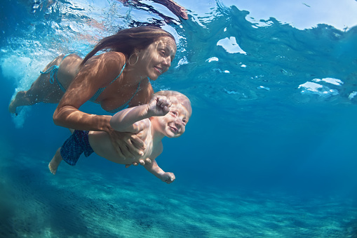 Happy family - mother hold in hands baby girl and dive down with fun in tropical beach sea pool. Active parent lifestyle, people water sport activity, underwater swimming lesson on vacation with child